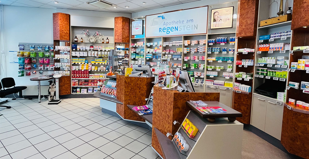 Heller Verkaufsraum einer Apotheke mit mehreren Beratungstheken, gut sortierten Regalen für Medikamente, Hautpflege- und Gesundheitsprodukte sowie einem zentralen Präsentationstisch.
Im Hintergrund sind weitere Produktregale und der Schriftzug „Apotheke am Regenstein“ zu sehen.