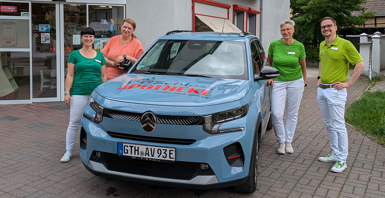 Vier Mitarbeitende der Schloss-Apotheke stehen vor dem Apothekeneingang und präsentieren ein hellblaues Elektroauto mit Apothekenlogo auf der Motorhaube.