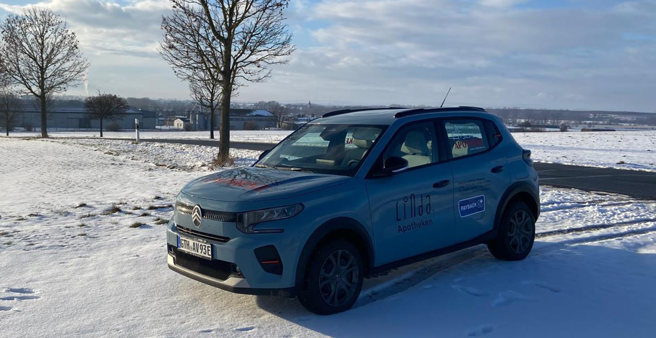 Hellblauer Citroën mit der Aufschrift ‚Linda Apotheken‘ steht auf einer verschneiten Landstraße. Die umliegenden Felder sind mit Schnee bedeckt; im Hintergrund stehen kahle Bäume, einige Gebäude und ein bewölkter Winterhimmel.