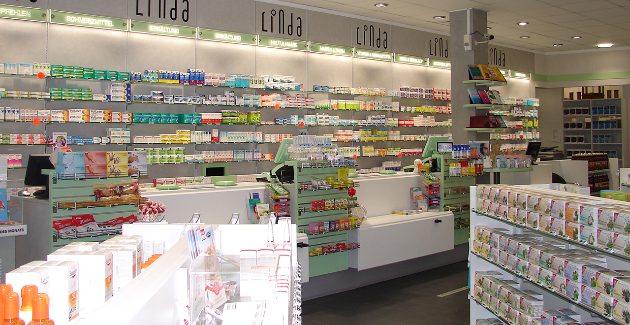 Panoramaaufnahme einer Linda-Apotheke mit einer langen Verkaufs- und Beratungsfläche. Die Wände sind mit Medikamentenregalen ausgestattet, beschriftet mit verschiedenen Gesundheitskategorien.
