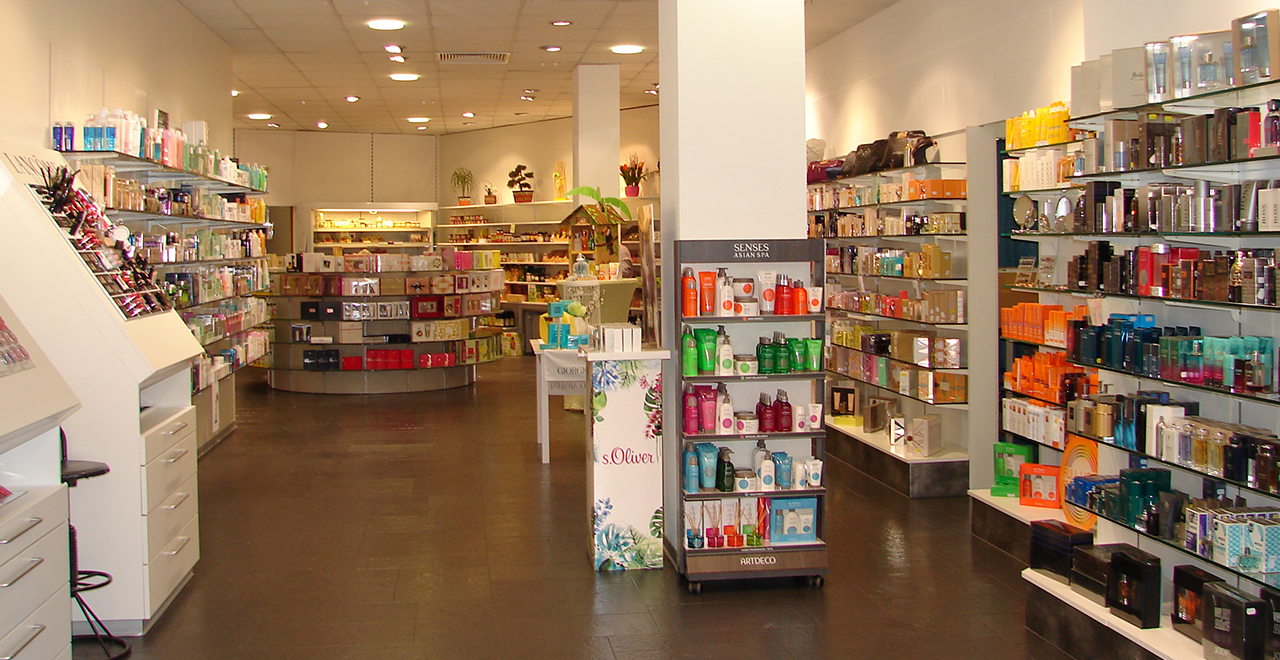 Weitläufiger Verkaufsbereich in einer Apotheke mit mehreren Regalen voller Kosmetikprodukte. In der Mitte steht ein Aufsteller mit Pflegeprodukten, links und rechts sind Regale mit Cremes, Shampoos und Parfüms. Der Raum ist hell und modern mit einer klaren Struktur.
