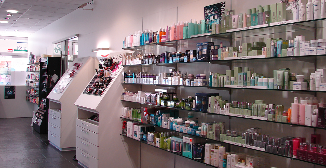 Lange Regalwand mit einer großen Auswahl an Hautpflegeprodukten und Make-up, sortiert nach Marken. Davor stehen zwei weiße Präsentationselemente mit dekorativen Kosmetikartikeln. Der Verkaufsbereich ist hell beleuchtet und klar gegliedert.