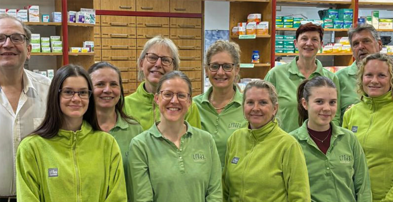 Gruppenfoto eines Apothekenteams in den Verkaufsräumen einer Apotheke. Insgesamt 13 Personen – Männer und Frauen – stehen lächelnd vor gut gefüllten Apothekenregalen. Die meisten tragen grüne Arbeitskleidung mit dem Linda-Apotheken-Logo. Im Hintergrund sind Medikamentenverpackungen und Holzkästen zur Aufbewahrung zu sehen.