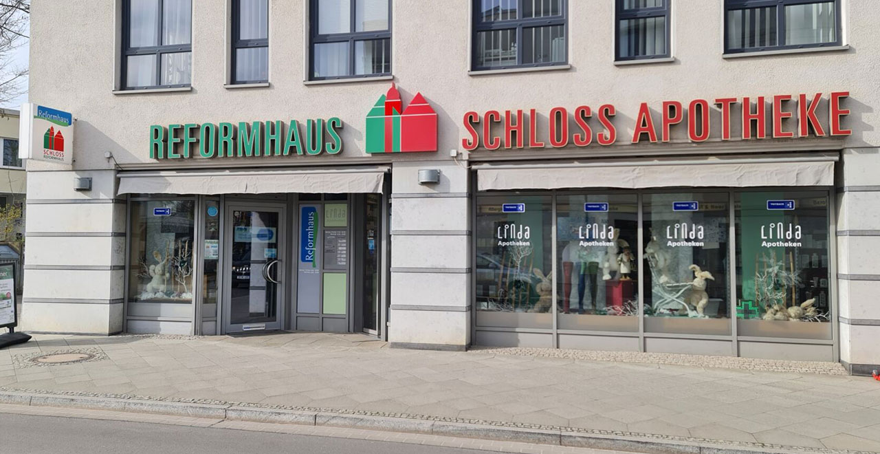 Eingangsbereich eines Gebäudes mit den Geschäften Reformhaus und Schloss Apotheke. 
Über den Schaufenstern befinden sich große grüne und rote Leuchtschriften. 
Vor dem Gebäude verläuft ein breiter Gehweg.