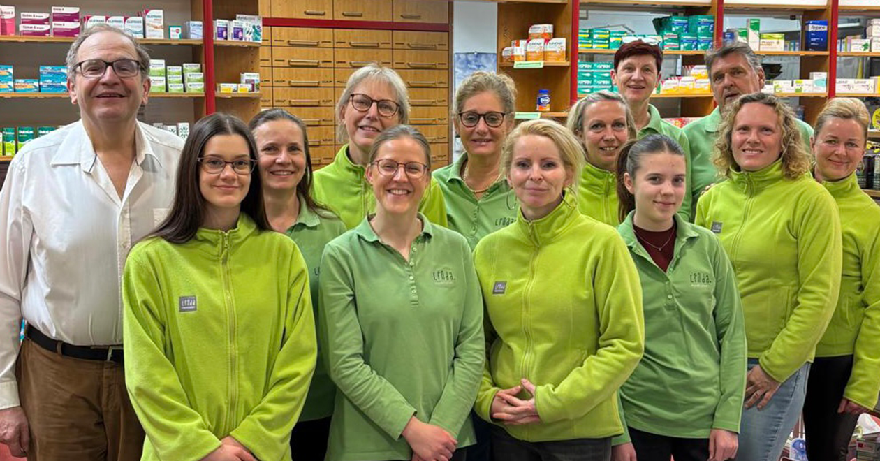 Gruppenfoto eines Apothekenteams in den Verkaufsräumen einer Apotheke. Insgesamt 13 Personen – Männer und Frauen – stehen lächelnd vor gut gefüllten Apothekenregalen. Die meisten tragen grüne Arbeitskleidung mit dem Linda-Apotheken-Logo. Im Hintergrund sind Medikamentenverpackungen und Holzkästen zur Aufbewahrung zu sehen.