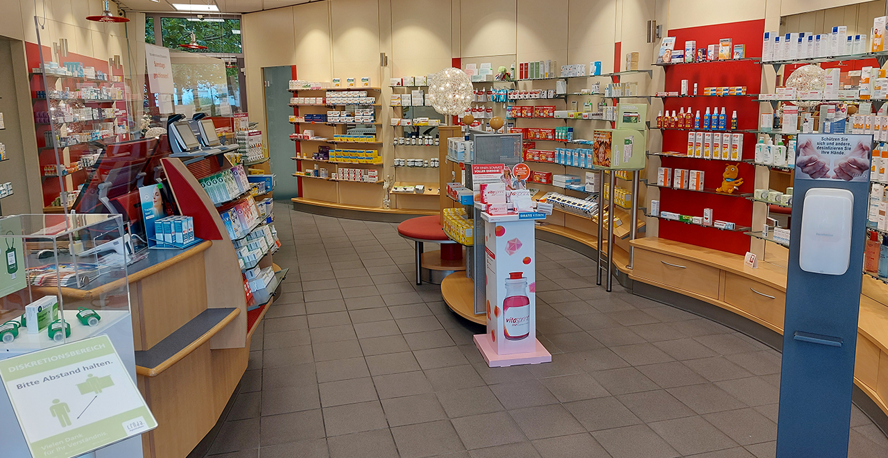 Blick in den Verkaufsraum der Apotheke mit modernen Regalen, Medikamenten und Hygieneprodukten. Eine runde Sitzgelegenheit steht in der Mitte.