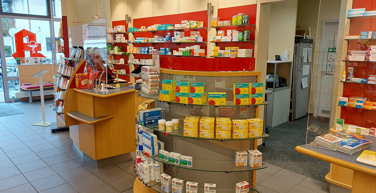 Weitere Innenansicht mit Verkaufsbereich, Kasse mit Plexiglasschutz und Regalen voller Arzneimittel. Helle und freundliche Einrichtung.