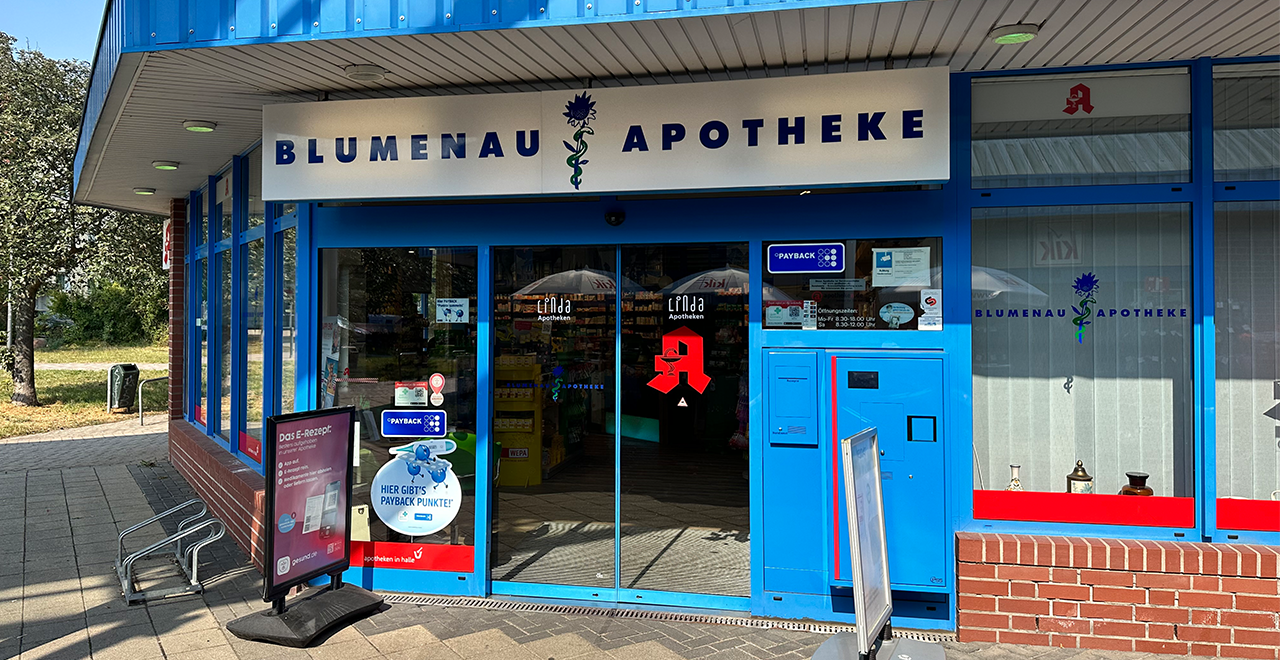 Frontansicht der „Blumenau Apotheke“ mit automatischen Glastüren und einem Schild über dem Eingang. Auf den Türen und Fenstern sind Apothekenlogos und Hinweise zur Kundenkarte (Payback) sichtbar. Direkt vor dem Eingang stehen ein Aufsteller mit Infos zum E-Rezept sowie ein blauer Automat. Links daneben ein Fahrradständer und ein Weg, der an einer Wiese vorbeiführt.