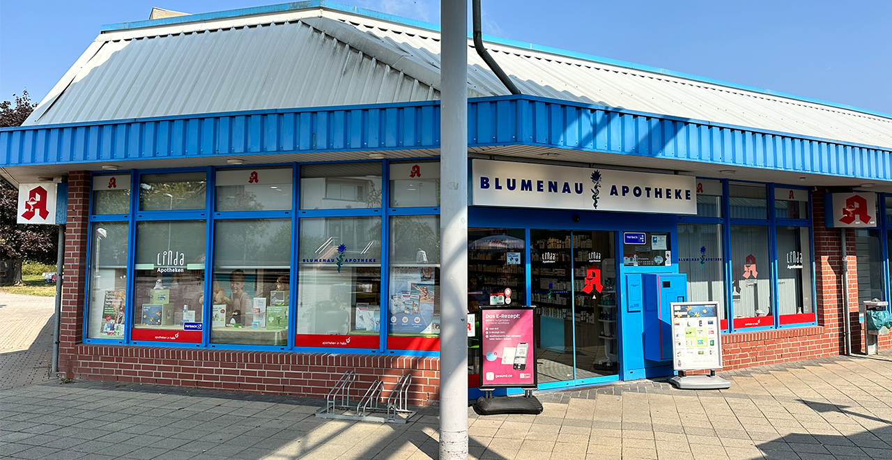 Außenansicht der „Blumenau Apotheke“ an einem sonnigen Tag. Das Gebäude hat eine blaue Fensterumrahmung und ein weißes Dach. Über dem Eingang ist ein Schild mit dem Apothekennamen angebracht. Vor dem Eingang stehen ein Fahrradständer und ein blauer Automat. In den Schaufenstern sind Apothekenzeichen und Werbeplakate zu sehen.