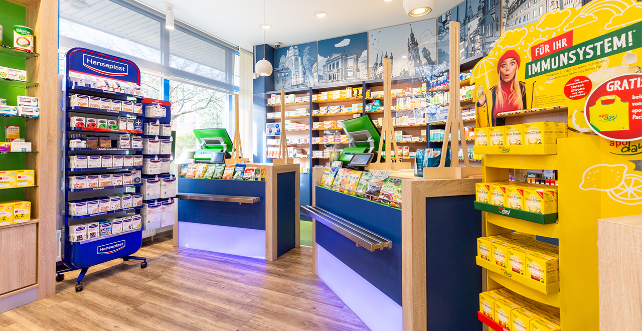 Blick auf den Kassenbereich einer Apotheke mit zwei modernen, grünen Kassensystemen. Links ein blaues Regal mit Hansaplast-Produkten. Rechts ein gelber Aufsteller mit dem Slogan „Für Ihr Immunsystem!“ und Vitaminprodukten. Die Rückwand zeigt eine Illustration von Stadtgebäuden.