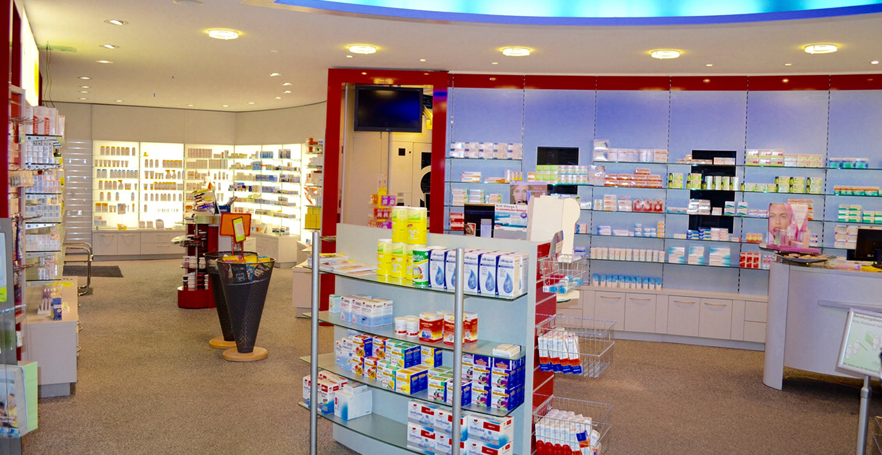 Innenansicht einer modernen Apotheke mit gut beleuchteten Regalen voller Medikamente, Pflegeprodukte und Nahrungsergänzungsmittel. Im Vordergrund befinden sich Verkaufsständer mit Sonderangeboten. Die Einrichtung ist hell, freundlich und übersichtlich gestaltet.