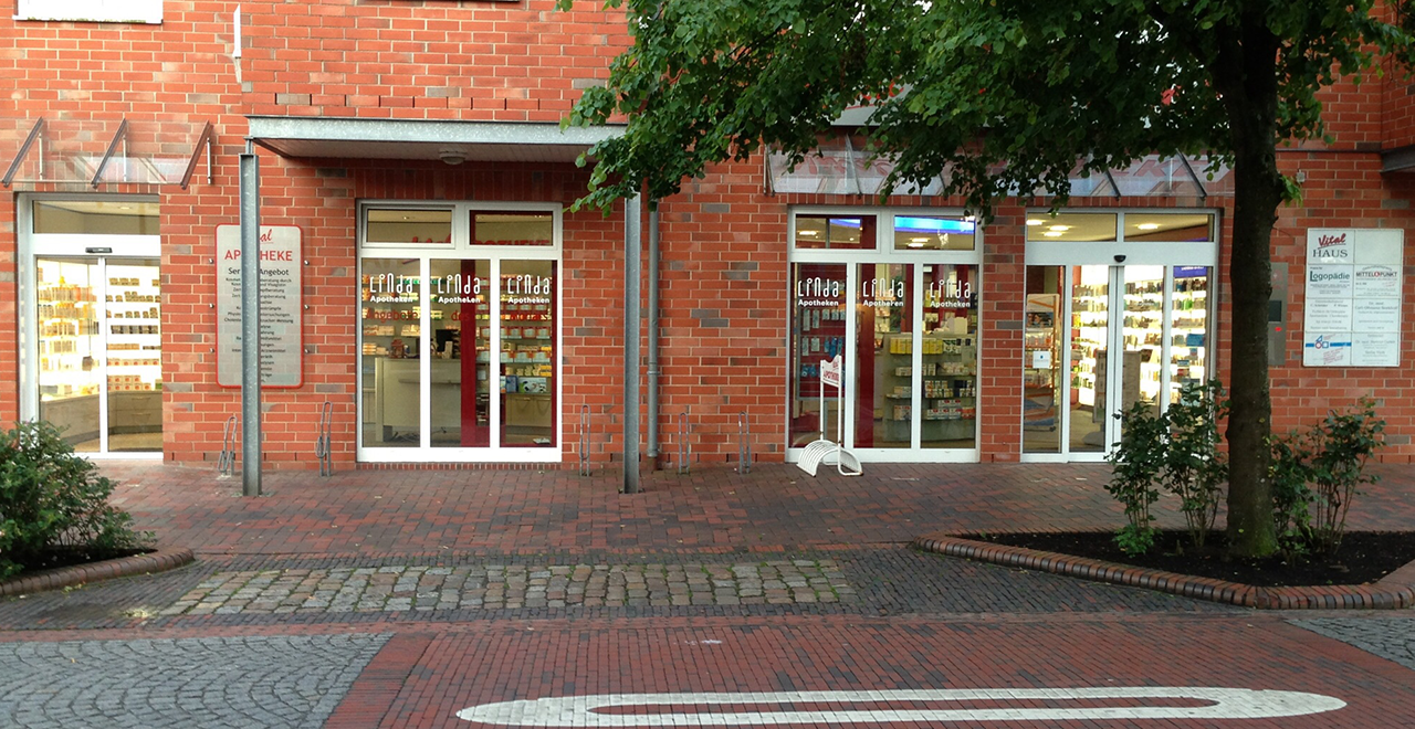 Außenansicht einer Apotheke in einem roten Backsteingebäude mit großen Schaufenstern und automatischen Glastüren. Über den Eingängen steht „Linda Apotheken“. Vor dem Gebäude befinden sich Pflastersteine, ein Baum und gepflegte Beete.