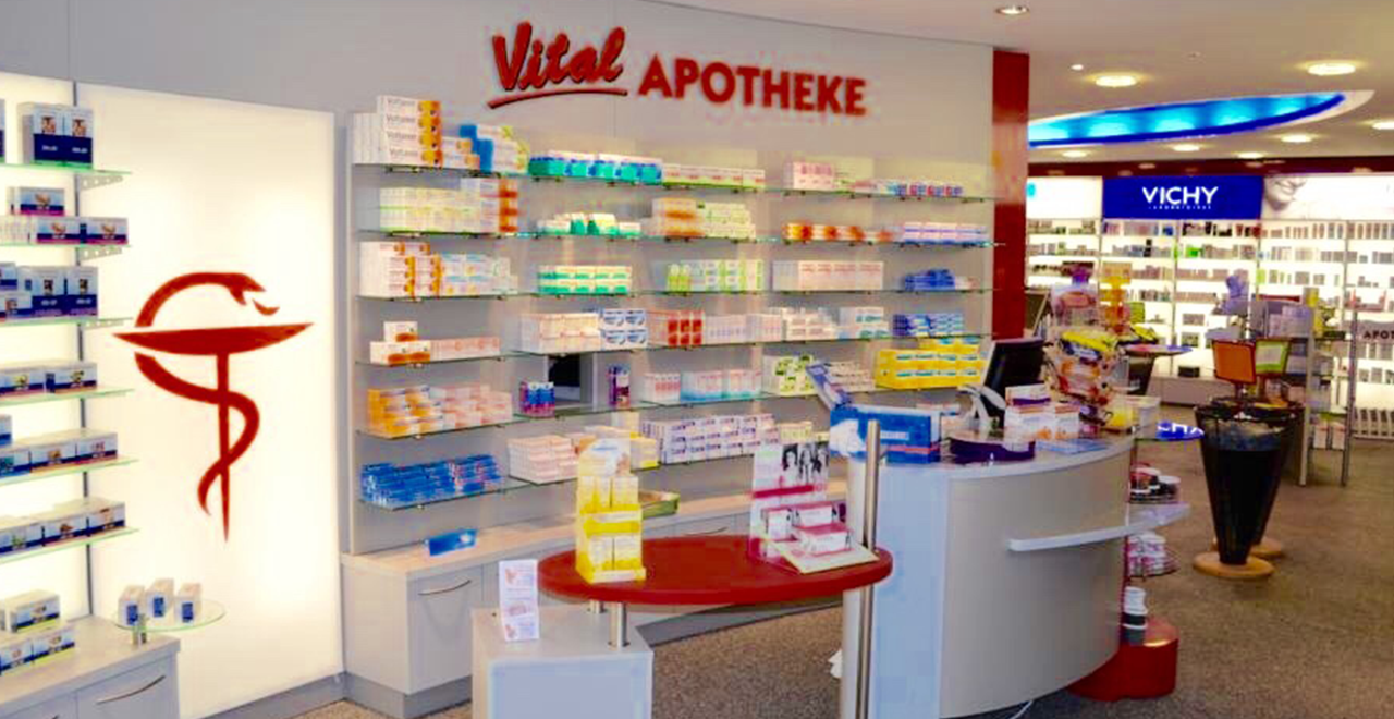 Blick auf den Verkaufsbereich einer Apotheke mit der Aufschrift „Vital Apotheke“. Die Rückwand ist mit Medikamenten und Produkten bestückt, links ist das rote Apothekenzeichen mit der Schlange sichtbar. Im Vordergrund stehen Theken mit weiteren Produkten.