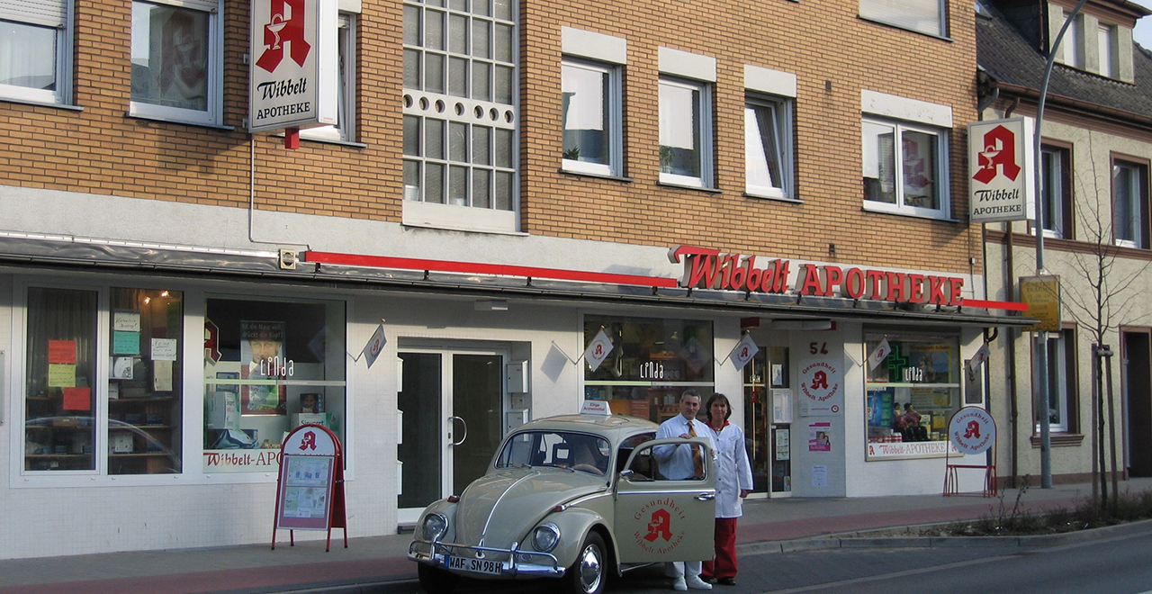 Außenansicht der Wibbelt Apotheke mit einem gelben VW Käfer vor dem Geschäft. Zwei Apotheker in weißen Kitteln stehen neben dem Auto. Das Schaufenster der Apotheke zeigt ein grünes Apothekenkreuz und verschiedene Werbeplakate. Über dem Eingang hängt ein großes Schild mit dem Namen „Wibbelt Apotheke“ in roten Buchstaben. Auf der rechten Seite ist ein weiteres Apothekenschild sichtbar.