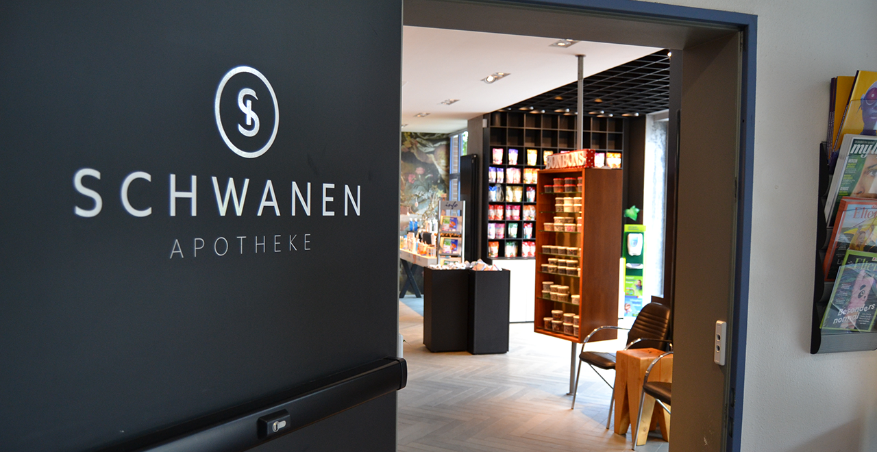 Eingang zur Schwanen Apotheke von innen. Links an der Wand steht das Apothekenlogo, durch die offene Tür ist der Verkaufsraum mit farbenfrohen Produkten und einem Bonbon-Regal sichtbar.
