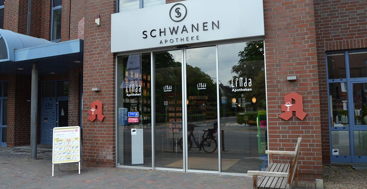 Außenansicht des Haupteingangs der Schwanen Apotheke mit automatischen Glas-Schiebetüren. Das Apothekenlogo und der Schriftzug „Linda Apotheken“ sind auf der Tür sichtbar. Ein Info-Aufsteller mit Angeboten steht links neben dem Eingang.