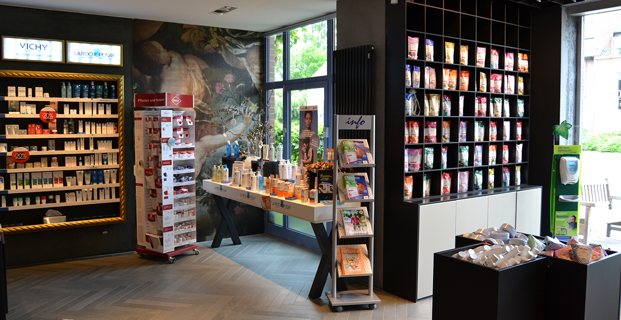 Verkaufsraum mit Schwerpunkt auf Kosmetik und Nahrungsergänzung. Im Bild sind ein Vichy- und La Roche-Posay-Regal, ein Werbetisch mit Pflegeprodukten, ein Magazin-Ständer und ein großes Regal mit Tütenprodukten zu sehen. Im Hintergrund ein Wandbild mit floralen Motiven.