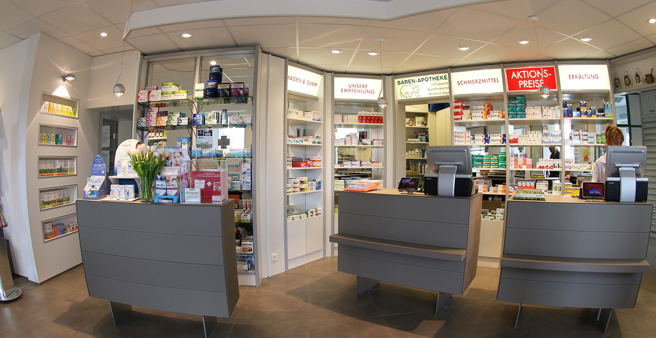 Heller Verkaufsraum einer Apotheke mit modernen grauen Theken, Regalen voller Medikamente und Kosmetikprodukte. Im Hintergrund sind beleuchtete Schilder mit Aufschriften wie „Schmerzmittel“, „Erkältung“ und „Aktionspreise“ zu sehen. Eine Mitarbeiterin steht hinter einer Kasse.