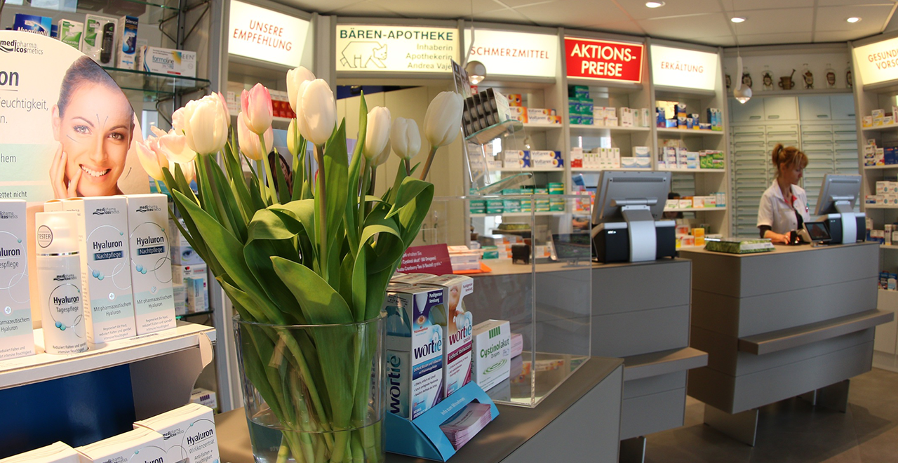 Nahaufnahme einer Apothekentheke mit einem Blumenstrauß aus weißen Tulpen und einer Produktpräsentation für Hyaluron-Pflegeprodukte. Im Hintergrund sieht man Apothekenmitarbeiterin an der Kasse und Regale mit Medikamenten.