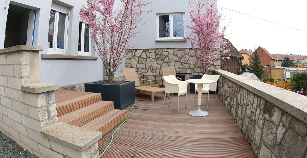 Außenbereich mit Holzterrasse, gemütlichen Sitzmöbeln und zwei blühenden Zierkirschen. Der Bereich ist von einer Steinmauer umgeben und lädt zum Verweilen ein. Im Hintergrund sind Nachbarhäuser sichtbar.