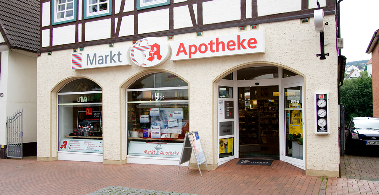 Vorderansicht der Markt-Apotheke in einem traditionellen Fachwerkhaus. Über den drei großen Schaufenstern und dem Eingang befindet sich das Apotheken-Schild mit der Aufschrift „Markt Apotheke“ sowie dem roten Apotheken-A. In den Schaufenstern sind Werbeplakate und Produkte ausgestellt, unter anderem von Linda Apotheken. Rechts neben dem Eingang befindet sich ein Wetter- und Uhrendisplay. Vor der Tür steht ein Kundenaufsteller mit einem Werbeplakat.