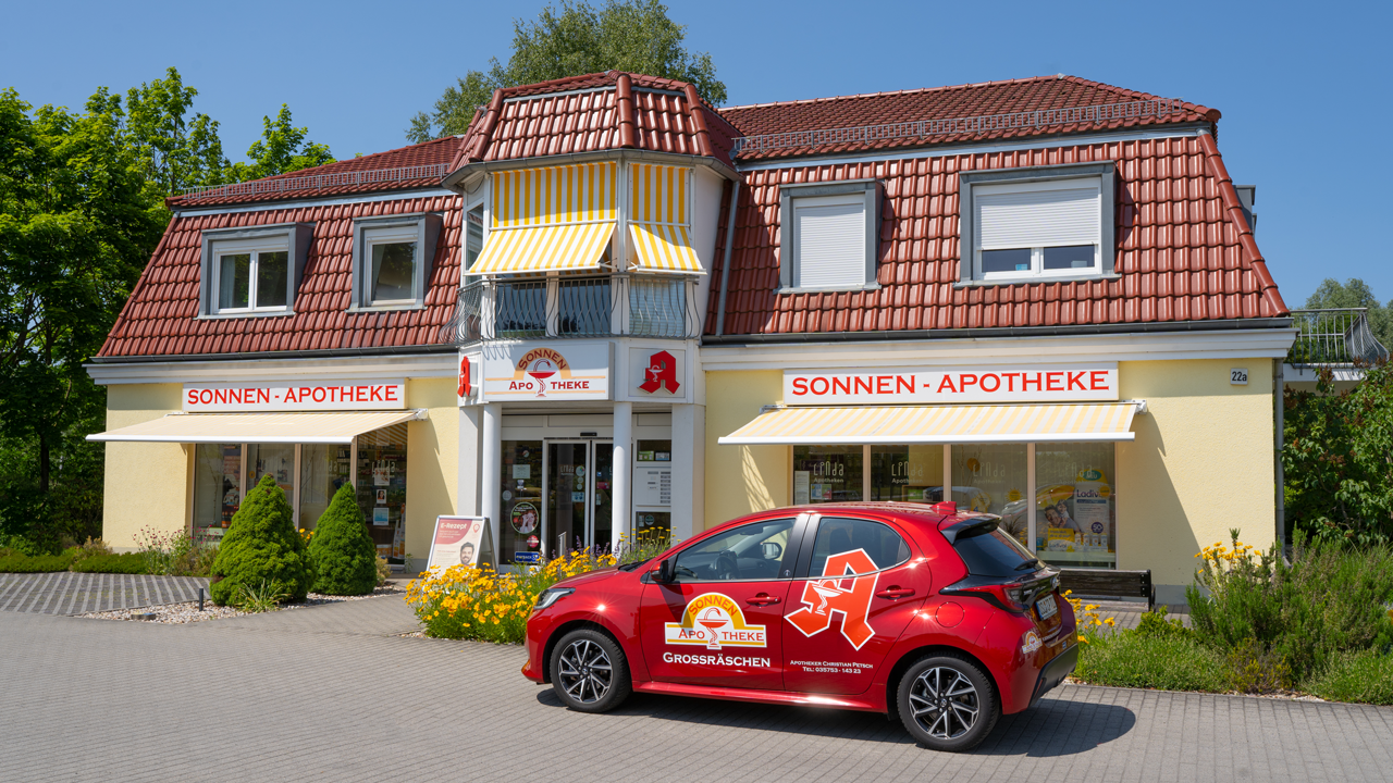 Gelbes Gebäude bei strahlend blauem Himmel, glänzendes Schild mit der Aufschrift Sonnenapotheke über der barrierefreien Eingangstür mit automatischen Schiebetüren. Beidseitig des Logos ist das rote Apotheken-A als Erkennungszeichen für öffentliche Apotheken angebracht. Links und rechts der Schiebetür befinden sich große Schaufenster mit geschmackvoller Dekoration, die von gelb-weiß gestreiften Markisen beschattet werden. Das rote, apothekeneigene Botendienstauto steht rechts auf dem Parkplatz vor einer gut gepflegten, naturnahen Rabatte.