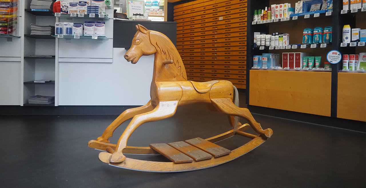 Ein hölzernes Schaukelpferd in einer Apotheke, das auf dem Boden in der Mitte des Raumes steht. Im Hintergrund sind Regale mit Medikamenten und Produkten sowie der Verkaufstresen zu sehen.