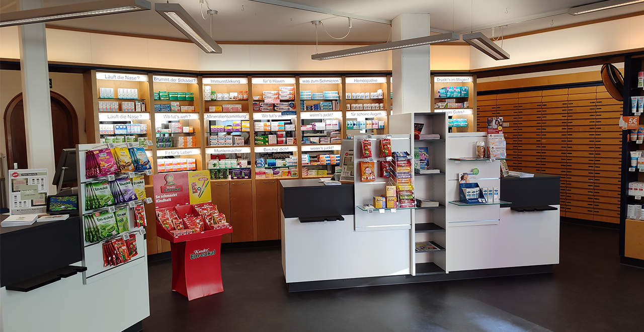 Eine Apotheke mit verschiedenen Regalen, die eine breite Auswahl an Medikamenten und Gesundheitsprodukten zeigen. Der Verkaufstresen steht im Vordergrund und es sind bunte Produkte, darunter Süßigkeiten und Nahrungsergänzungsmittel, sichtbar.