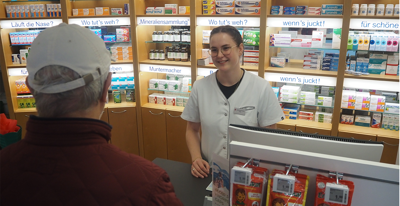 Eine Apothekerin steht hinter dem Verkaufstresen einer Apotheke und berät freundlich einen älteren Kunden. Im Hintergrund sind beleuchtete Regale mit verschiedenen Medikamenten und Produkten sichtbar, die thematisch beschriftet sind – zum Beispiel mit „Läuft die Nase?“ oder „wenn’s juckt!“. Die Atmosphäre wirkt einladend und professionell.
