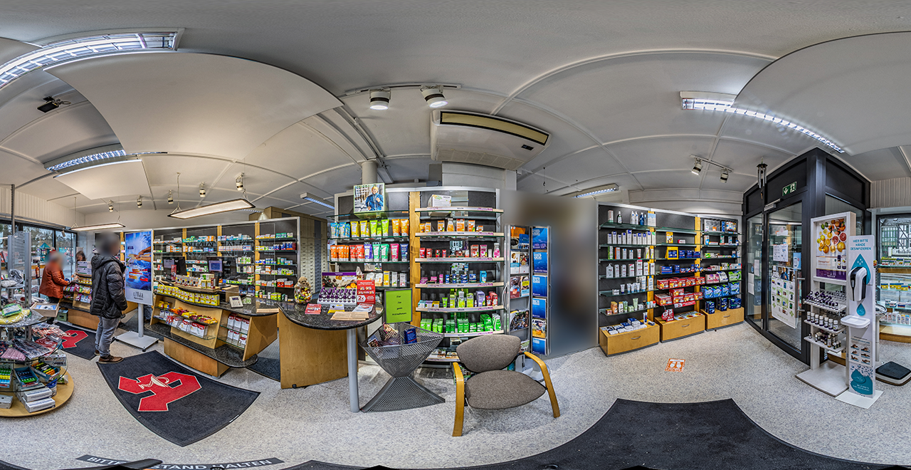 Innenansicht einer modernen Apotheke mit mehreren Regalen voller Kosmetik-, Pflege- und Gesundheitsprodukte. Zwei Personen stehen an der Kasse. Im Vordergrund befindet sich ein runder Tisch mit Werbeprospekten und ein grauer Stuhl. Die Decke ist gewölbt und mit hellen LED-Leuchten ausgestattet.