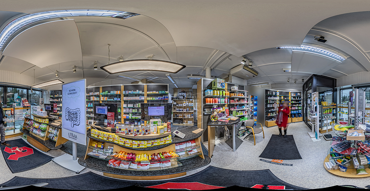 Panorama-Ansicht einer Apotheke mit zentralem Verkaufstresen, auf dem sich zahlreiche Medikamente und Nahrungsergänzungsmittel befinden. Links steht ein digitales Werbedisplay mit einer Darmgrafik und dem Text „Schon gewusst?“. Im rechten Bereich geht eine Frau in rotem Mantel durch den Laden.