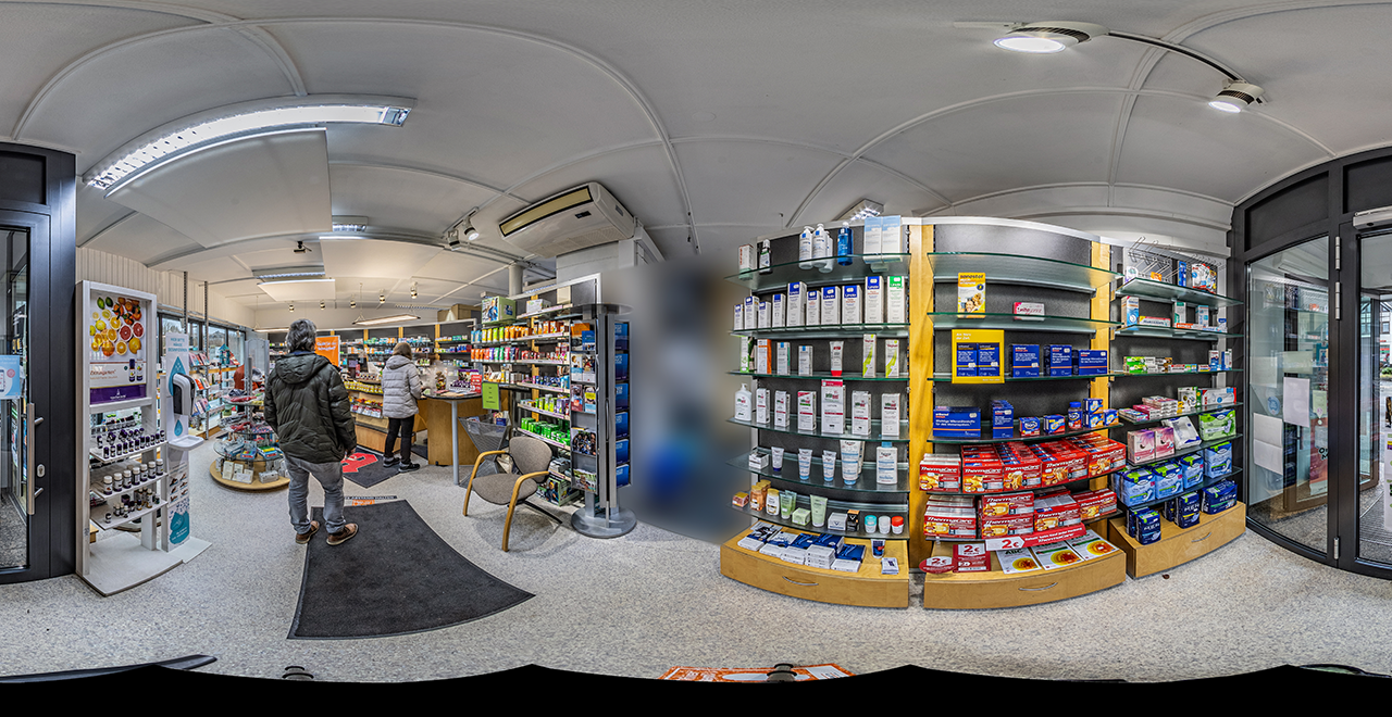 Blick von einem Eingang in eine Apotheke mit mehreren Regalen voller Gesundheitsprodukte, Arzneimittel und Pflegeartikel. Ein Mann in Winterjacke steht an einem schwarzen Teppich mit Abstandsmarkierung. Links ein runder Präsentationstisch mit Produkten, rechts Regale mit Schmerzmitteln und Hautpflege.