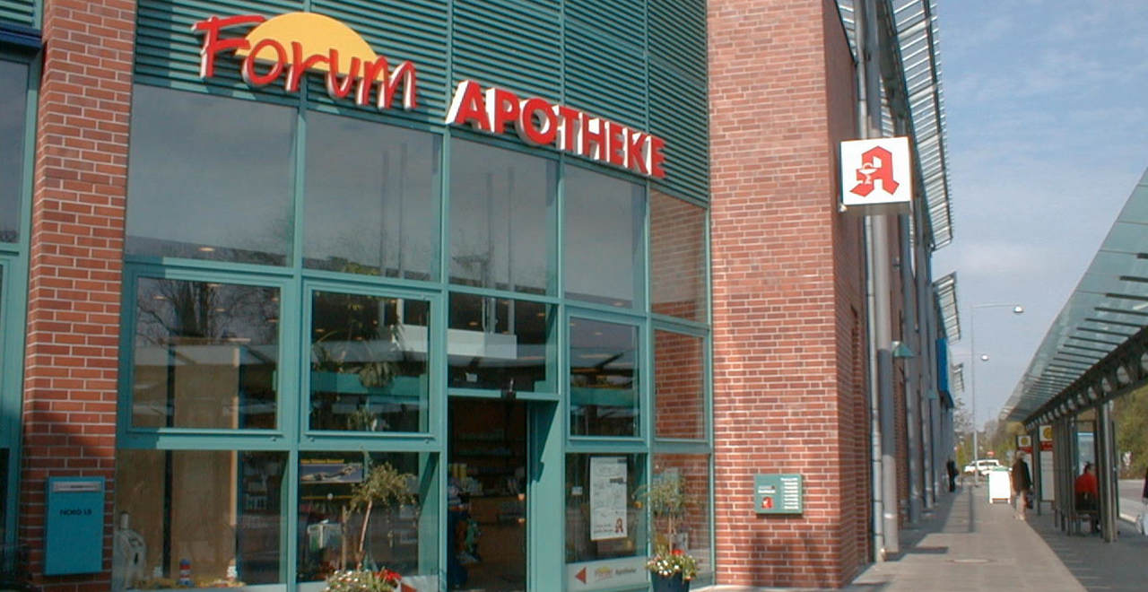 Das Foto zeigt die Außenansicht einer Apotheke mit der Aufschrift 'Forum Apotheke' in großen roten Buchstaben. Über der Eingangstür ist ein großes gelbes Symbol zu sehen, das eine Sonne darstellt. Der Eingang ist gut sichtbar, und es gibt eine rote Apotheken-Symbolbeleuchtung an der Wand. Die Fenster sind groß und der Eingang ist gut zugänglich.