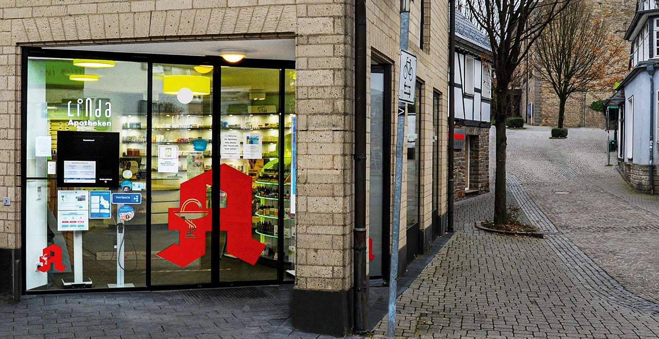Eingang einer Linda Apotheke an einer gepflasterten Gasse in einer Altstadt mit Fachwerkhäusern. Die Apotheke befindet sich in einem modernen Gebäude mit heller Klinkerfassade. Durch die großen Glasfenster sind Regale mit Produkten sichtbar. Auf der Tür ist das rote Apotheken-A gut erkennbar. Rechts vom Eingang führt eine ruhige Gasse leicht bergauf, gesäumt von Bäumen und traditionellen Gebäuden.
