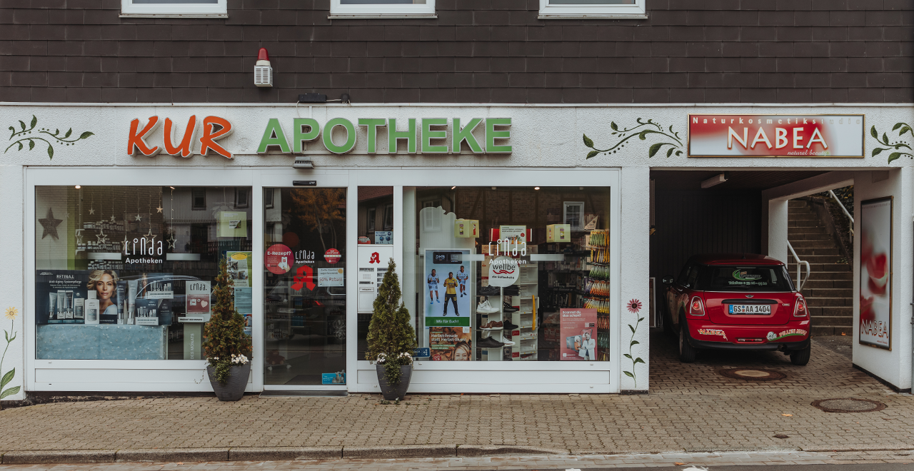 Straßenansicht einer Apotheke mit der groß beschrifteten Fassade „KUR APOTHEKE“. Die Eingangstür besteht aus breiten Glasschiebetüren, durch die man Regale und Werbeplakate im Inneren erkennt. Links und rechts des Eingangs stehen zwei Pflanzenkübel. Direkt neben dem Eingang befindet sich eine Durchfahrt mit einem roten Auto darin und einem Schild „NABEA Naturkosmetikstudio“ über der Einfahrt. Die Hauswand ist weiß und mit Blumenmotiven verziert.