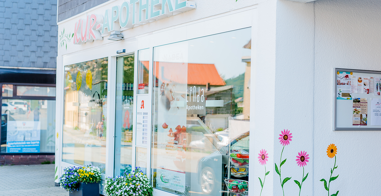 Glasfront der Kur-Apotheke mit Blumendekoration und Blick ins Innere mit Produkten im Schaufenster.