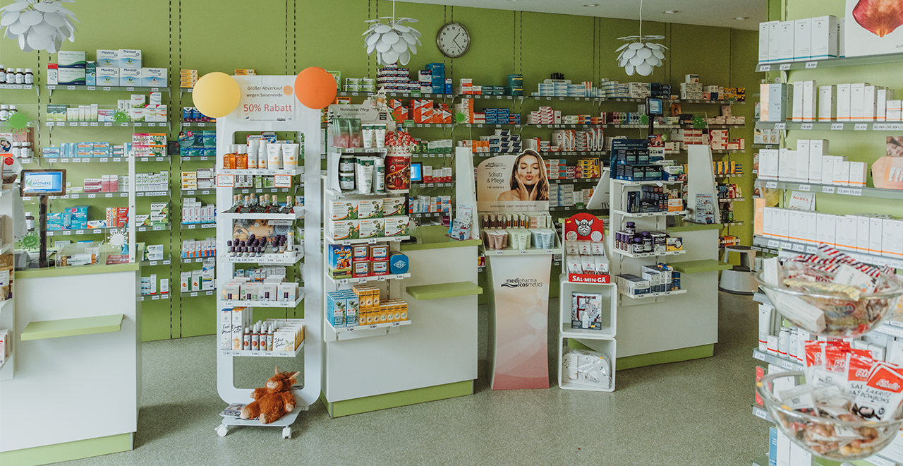 Ein weiterer Blick in dieselbe Apotheke: Im Vordergrund stehen mehrere weiße Präsentationsmodule mit Pflegeprodukten, Nahrungsergänzungsmitteln und einem Werbeaufsteller mit dem Gesicht einer Frau. Oben hängen dekorative Deckenlampen in Blütenform. Dahinter sind hohe Regale voller Arzneimittelpackungen, sortiert nach Farben und Größen. Links und rechts des Bildes stehen zusätzliche Regale mit Kosmetik- und Gesundheitsartikeln.