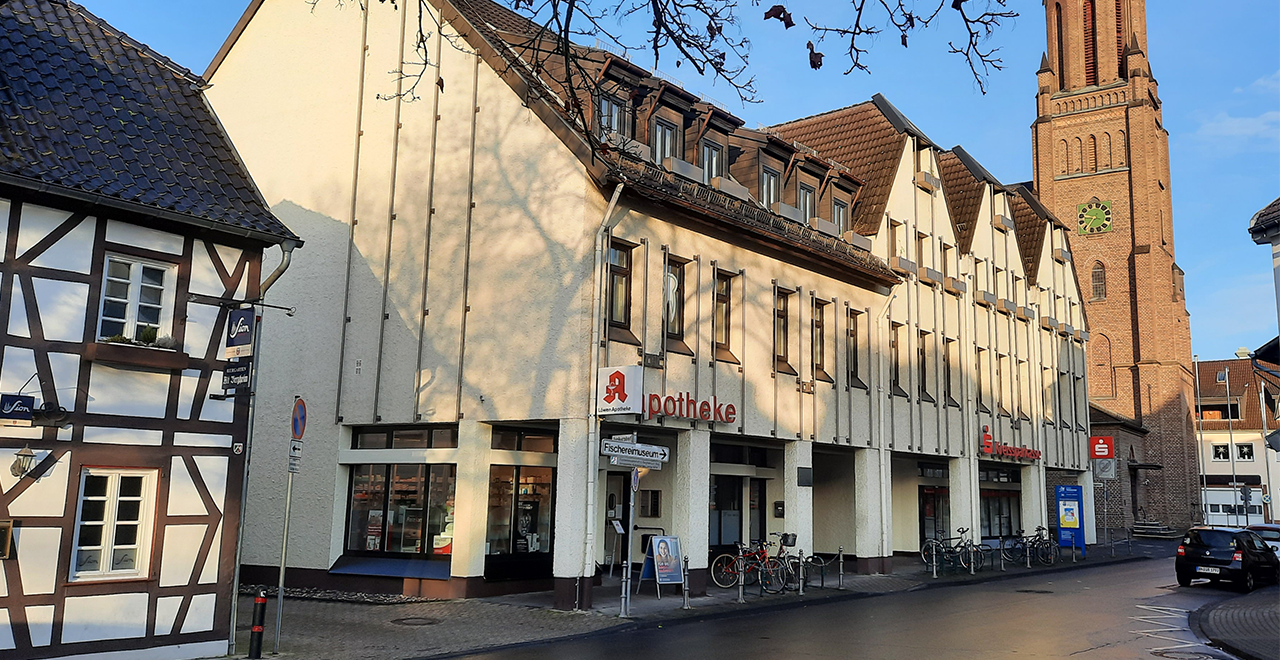 Eine Apotheke an einer Straße mit Fachwerkhaus links und Kirchturm im Hintergrund. Das Apotheken-Schild mit rotem Logo ist gut sichtbar.