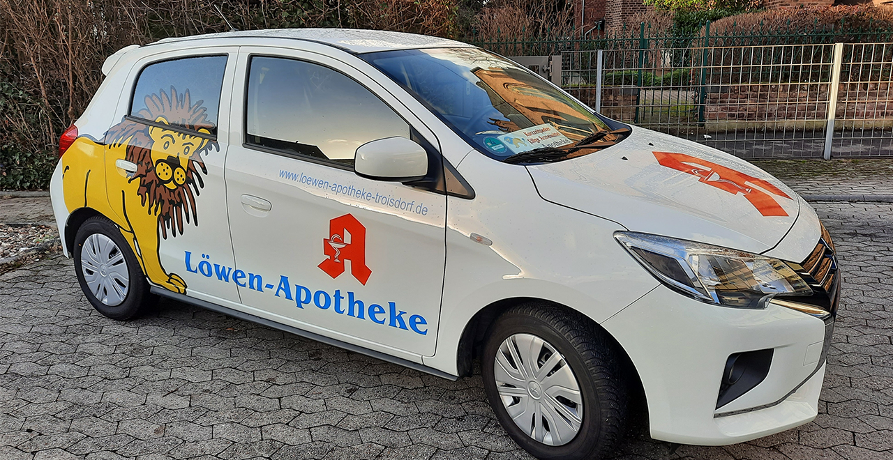 Ein weißes Auto mit Löwenzeichnung und der Aufschrift „Löwen-Apotheke“. Auf dem Auto sind das Apotheken-Logo und eine Webadresse zu sehen.