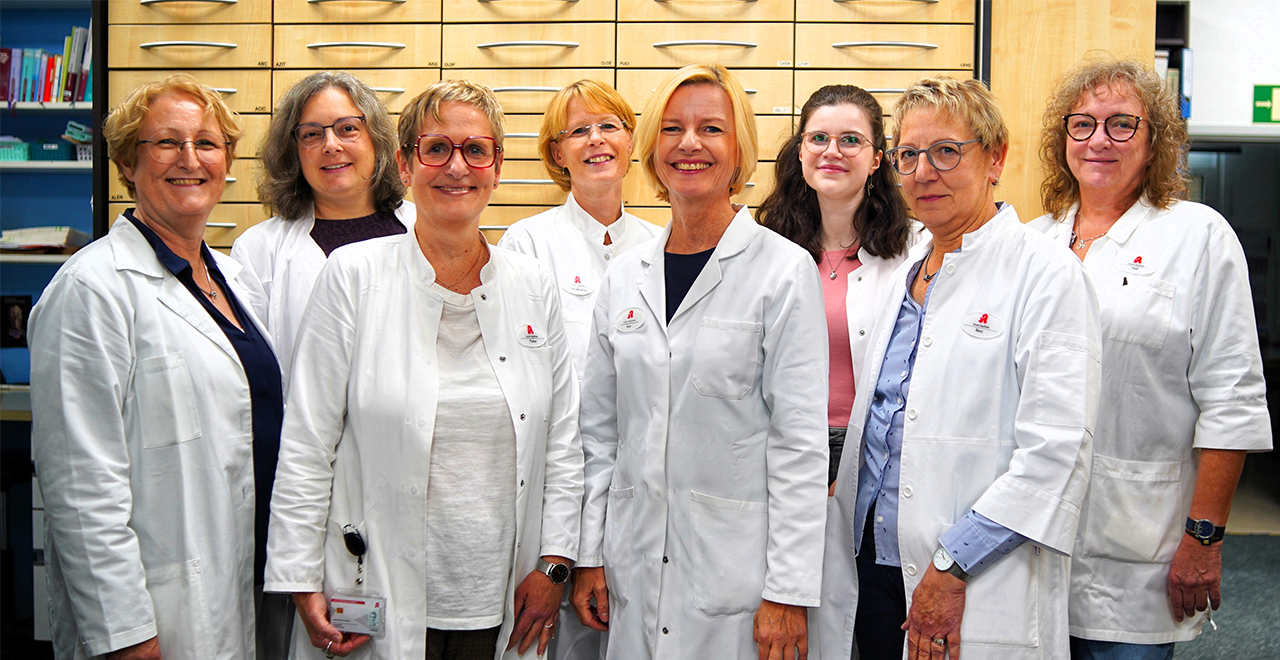 Acht Frauen in weißen Apothekenkitteln stehen in einer Apotheke und lächeln in die Kamera.