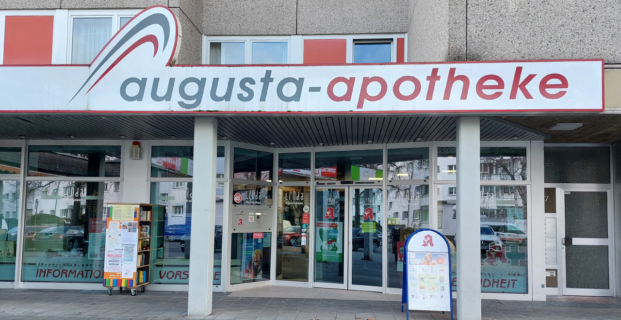 Das Bild zeigt die Außenansicht der Augusta-Apotheke mit einem großen roten und weißen Schild. Der Eingangsbereich ist gut sichtbar, mit einem Rollwagen, der verschiedene Flyer und Informationen über Gesundheit und Apotheke zeigt. Hinter den Fenstern sind Regale und Schilder sichtbar, die auf Angebote und Dienstleistungen hinweisen.