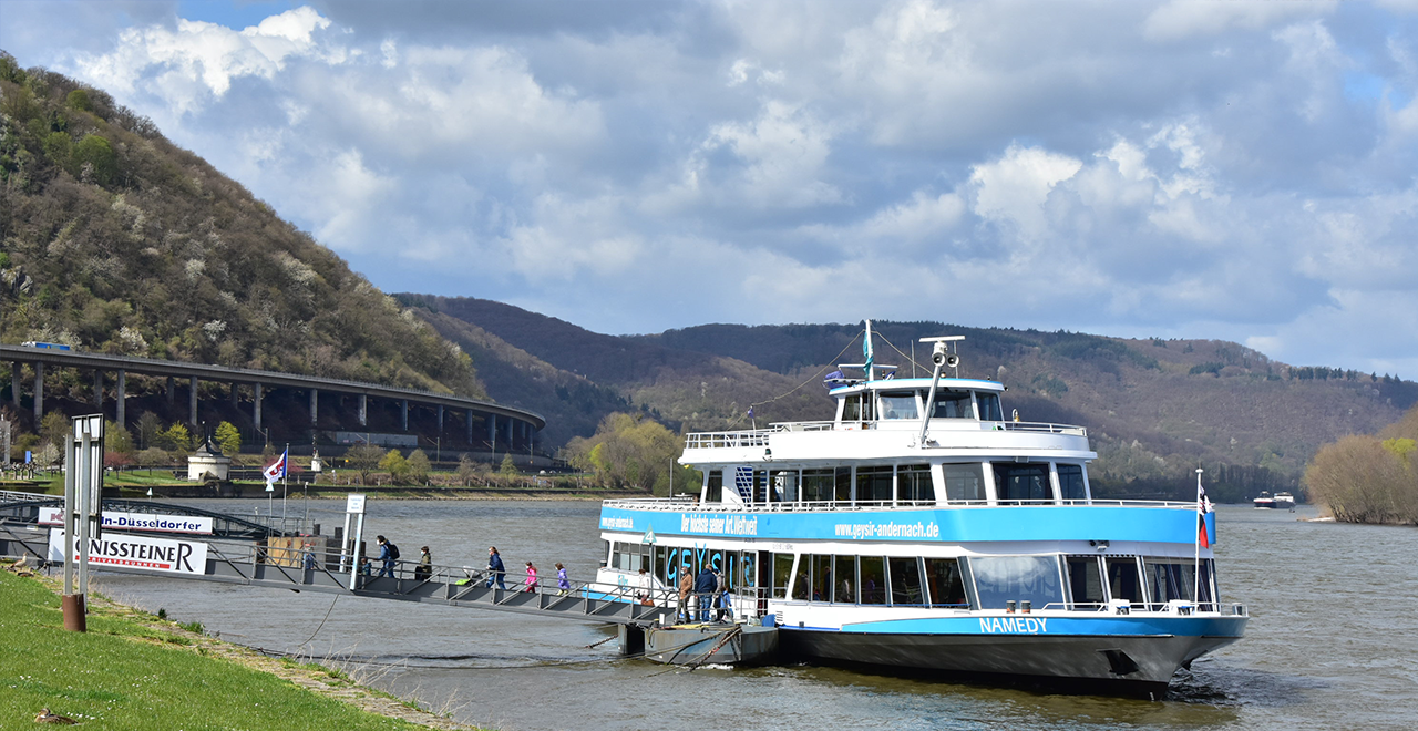 Passagierschiff 'NAMEDY' legt am Rheinufer bei Andernach an, im Hintergrund bewaldete Hügel und eine Straßenbrücke.