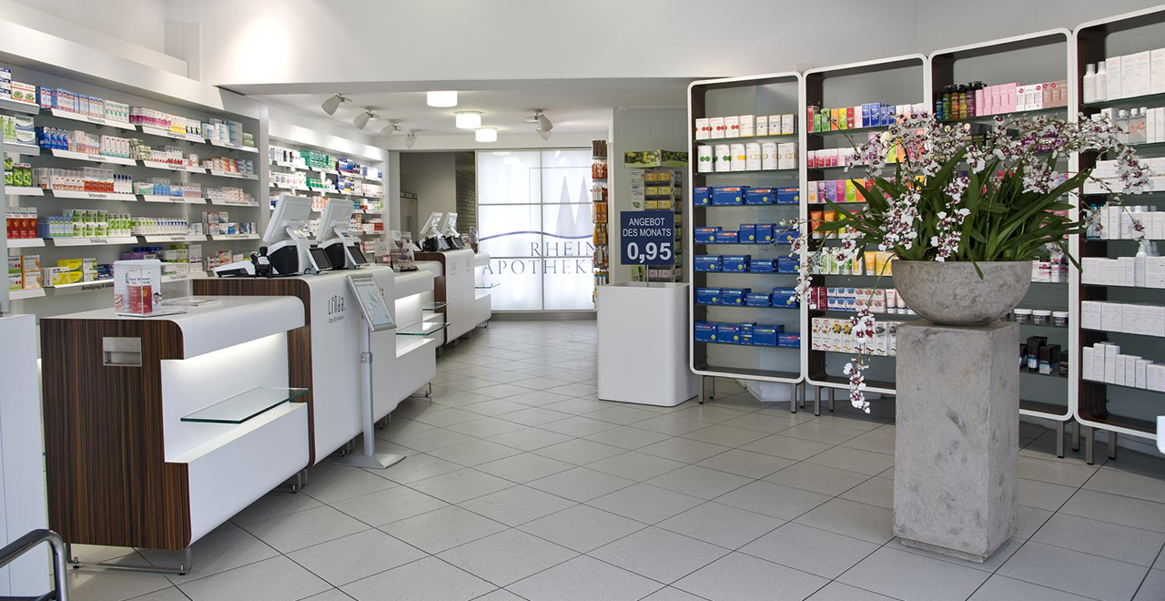 Innenraum einer modernen LINDA Apotheke mit zentralem Blumenarrangement, Verkaufsregalen und Theken mit Kassensystem. Im Hintergrund eine leuchtende Trennwand mit Apotheken-Logo.
