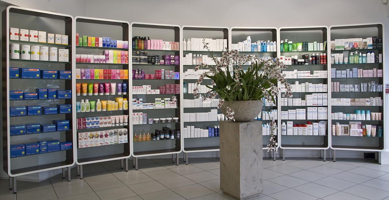 Große Regalwand mit farblich sortierten Pflegeprodukten und Medikamenten. Davor steht ein hoher Stein-Blumentopf mit weißen Orchideen.