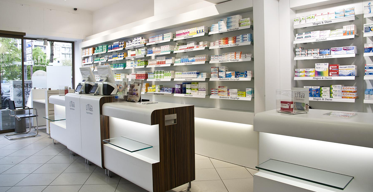 Innenansicht mit Blick zum Eingang der Apotheke. Links Glastür, rechts Verkaufsregale mit medizinischer Hautpflege. Moderne Theken und Kassensysteme sind mittig platziert.
