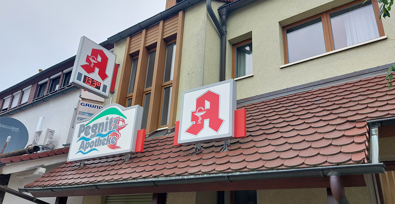 Außenansicht der Pegnitz Apotheke mit rotem Apothekenzeichen und digitaler Uhr.