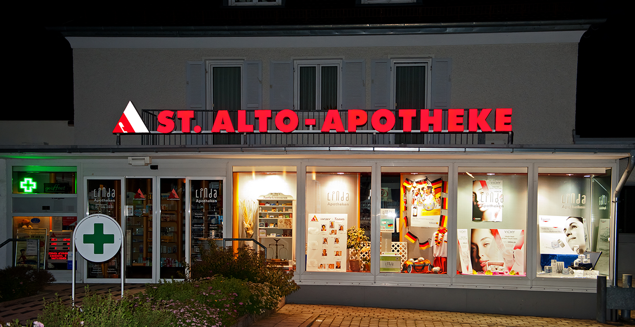 Beleuchtete Außenansicht der St. Alto Apotheke bei Nacht mit rotem Schriftzug und grünem Apothekenkreuz.