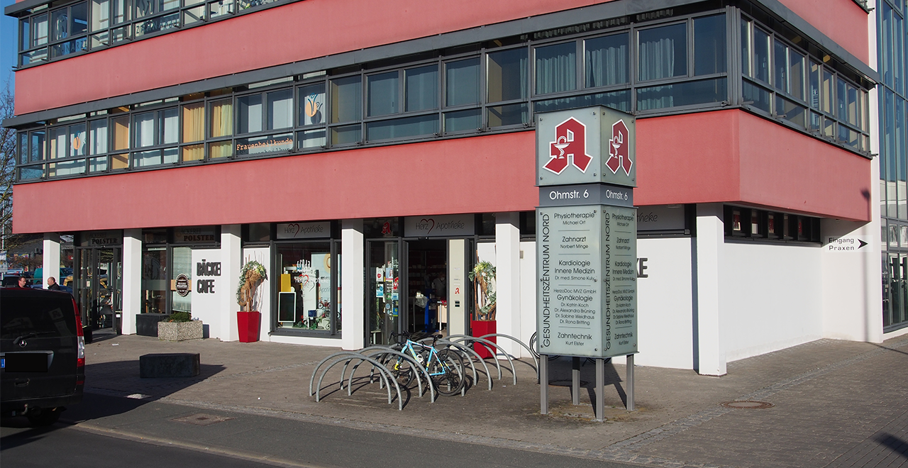 Modernes, barrierefreies Gebäude mit rot-orangener Fassade und großen Fensterfronten.
Im Erdgeschoss befinden sich mehrere Geschäfte und Praxen, darunter auch die Herz Apotheke.
Vor dem Eingang steht ein freistehendes Hinweisschild mit rotem Apotheken-Symbol und der Aufschrift „Ohmstr. 6“ sowie einer Liste medizinischer Einrichtungen.
Daneben gibt es Fahrradständer mit abgestellten Fahrrädern.