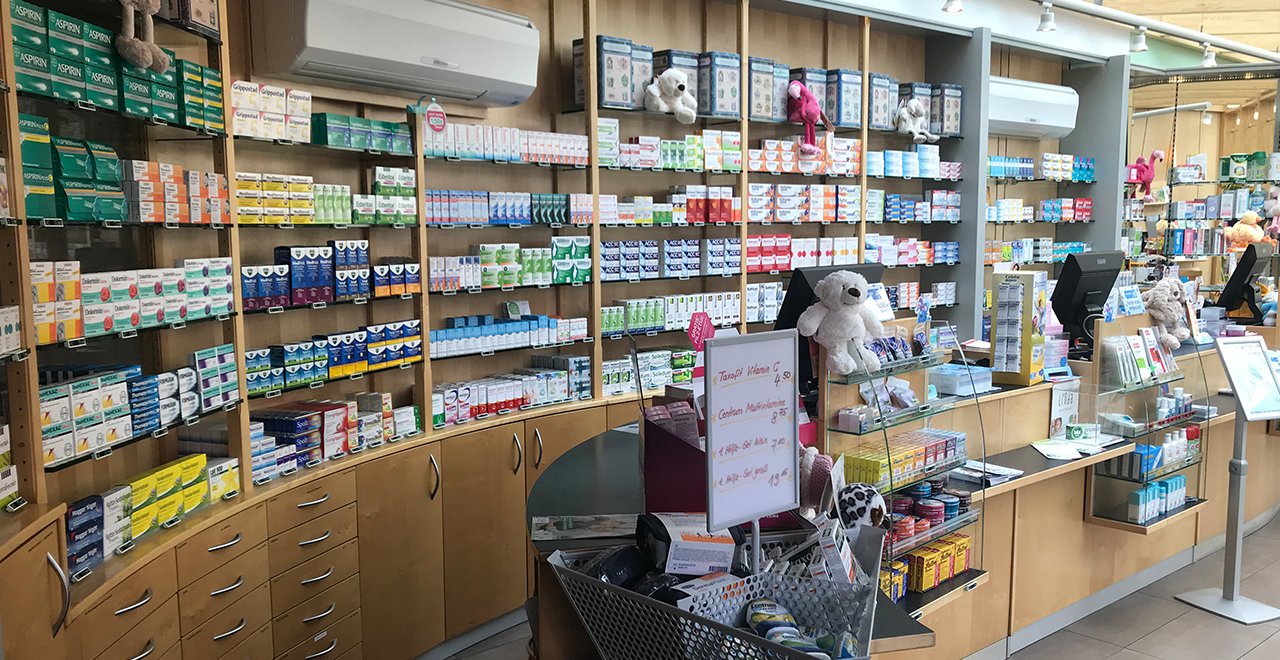 Innenansicht einer Apotheke mit Regalen voller Arzneimittel und Pflegeprodukte. In der Mitte des Bildes ist der Verkaufstresen mit einer Theke zu sehen, auf der Produkte ausgestellt sind. An den Wänden sind viele Arzneimittel in verschiedenen Verpackungen aufgereiht. Plüschtiere, darunter ein Bär, schmücken den Bereich hinter der Theke. Am Rand des Bildes sind zwei Computerarbeitsplätze zu erkennen.