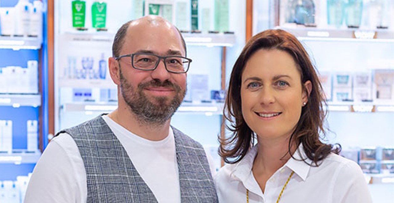 Das Bild zeigt den Inhaber einer Apotheke zusammen mit seiner Frau, die beide freundlich in die Kamera blicken. Sie stehen inmitten eines Apothekenregals mit verschiedenen Kosmetikprodukten und Gesundheitsartikeln im Hintergrund. Beide tragen gepflegte, moderne Kleidung und strahlen Professionalität und Freundlichkeit aus.